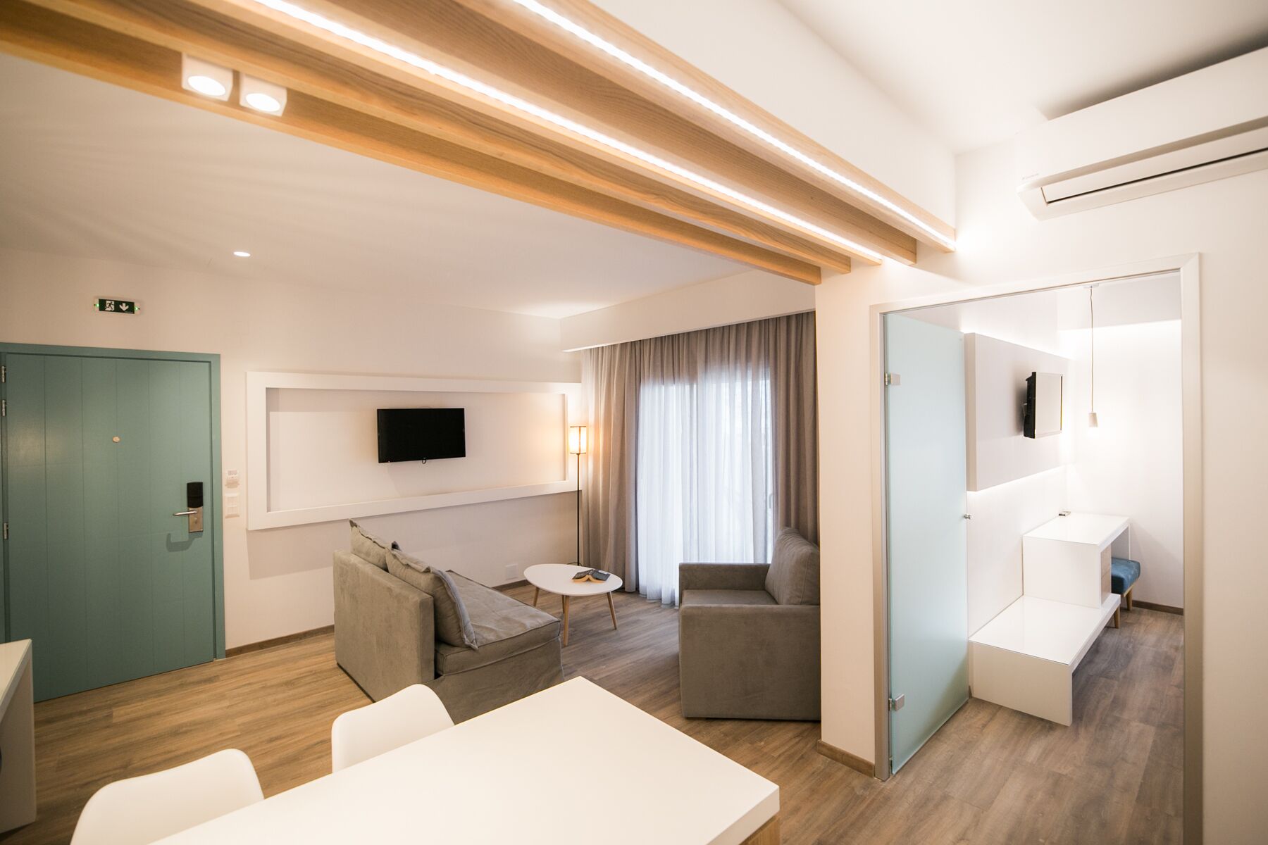 Een moderne hotelkamer met een houten plafond, een flatscreen televisie aan de muur, een bank, een klein rond tafeltje en een lichtgekleurde vloer.
