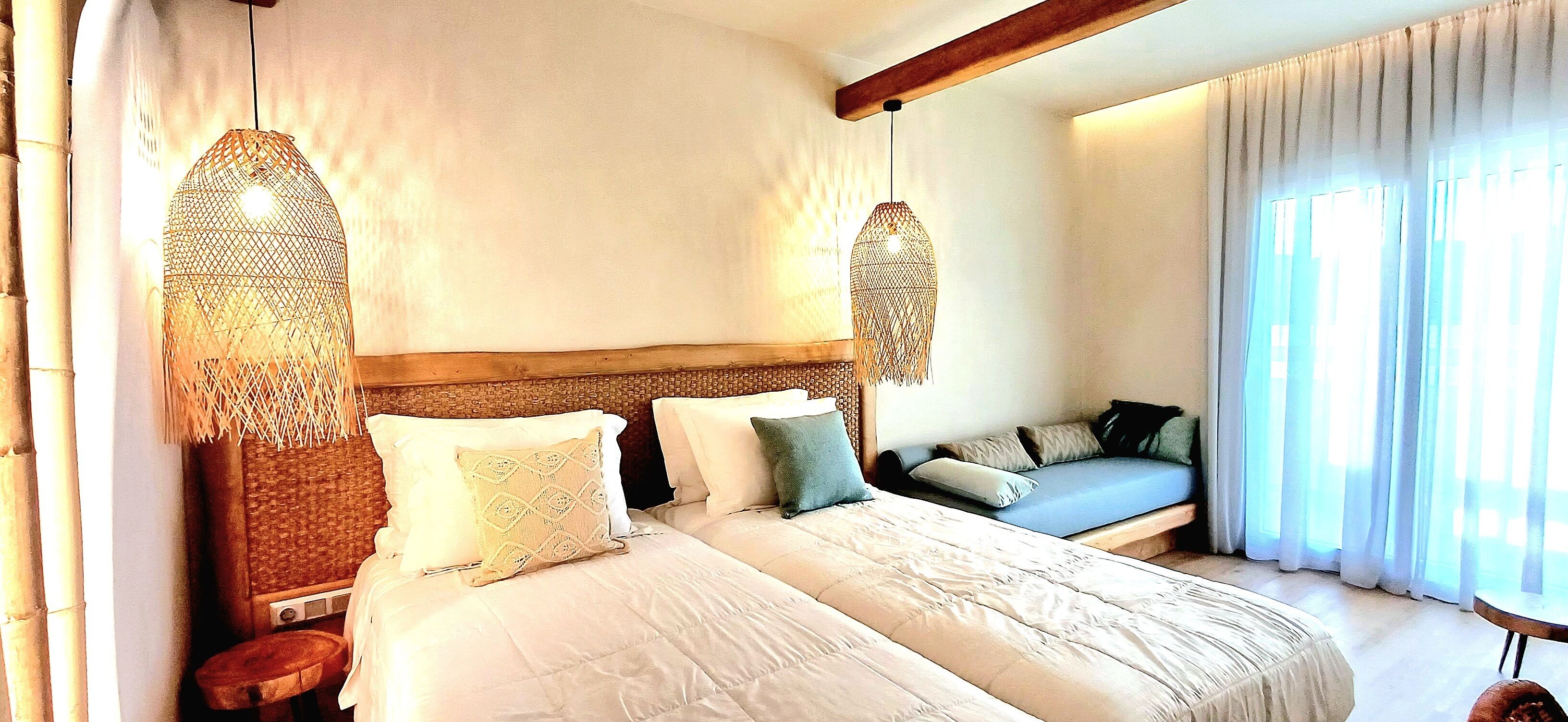 Quarto de hotel com uma cama grande, um sofá e uma janela com cortinas translúcidas.