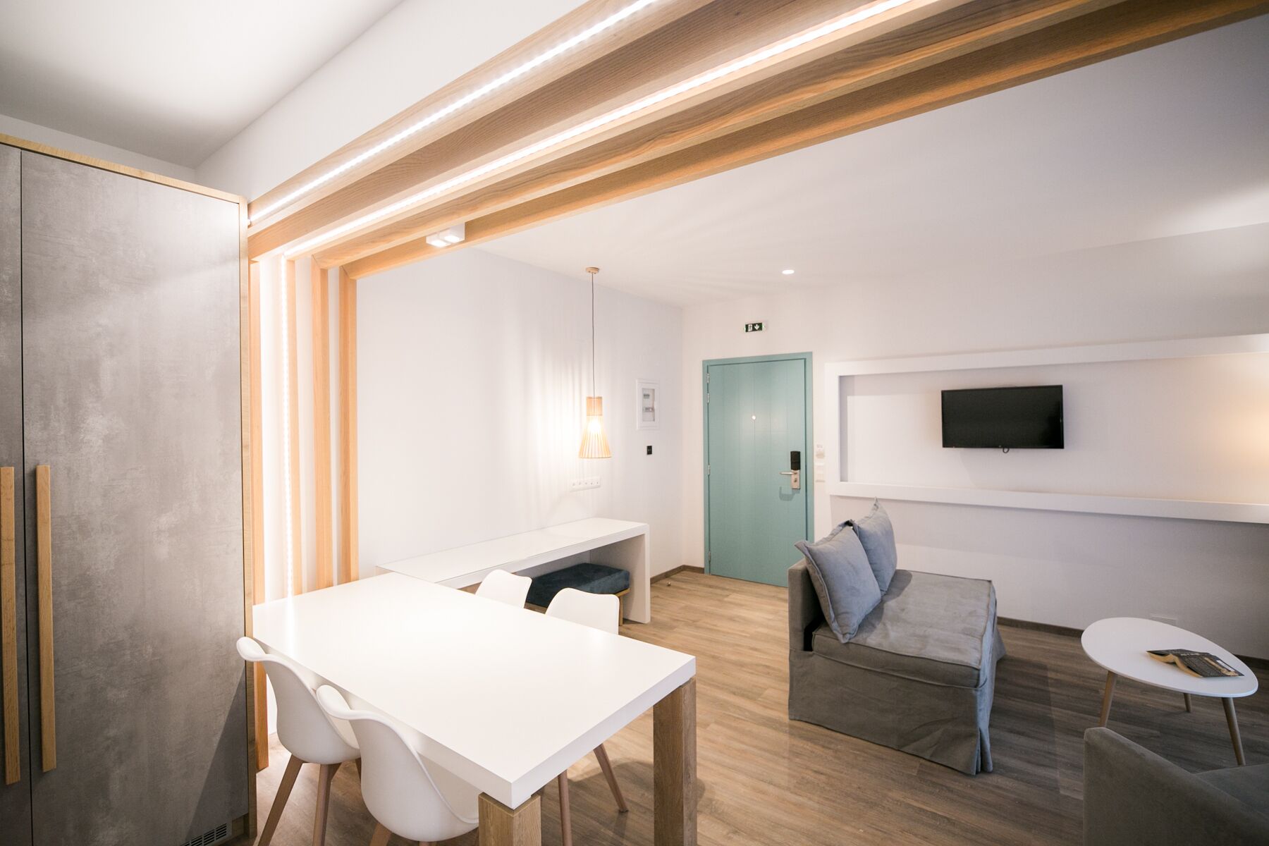 A modern hotel room with a white desk, chairs, a sofa, a small round table, and a TV mounted on the wall.