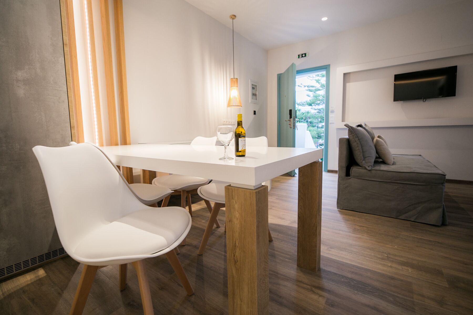 Ein moderner Essbereich mit einem weißen Tisch, Holzstühlen und einer Pendelleuchte. Auf dem Tisch stehen eine Weinflasche und ein Glas.