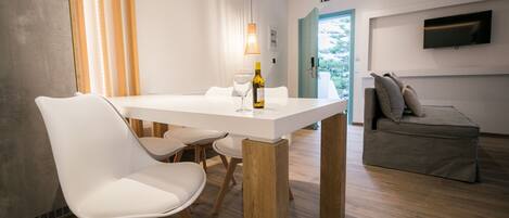 A modern dining area with a white table, wooden chairs, and a pendant light. A bottle of wine and a glass are on the table.