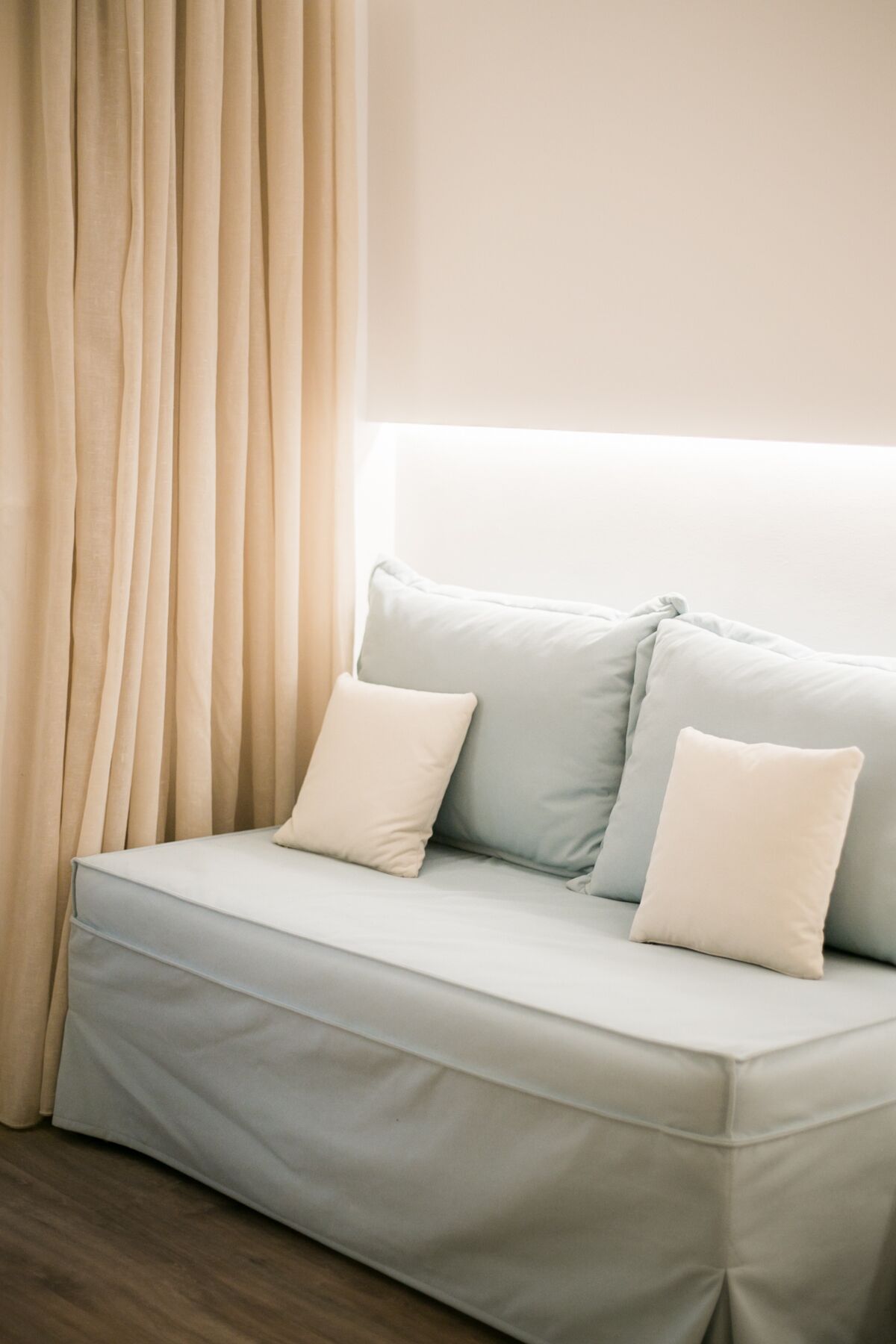 Un sofá de color claro con dos almohadas apoyado contra una pared, una ventana con cortinas y un suelo de madera.