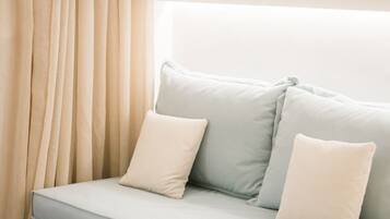 Un sofá de color claro con dos almohadas apoyado contra una pared, una ventana con cortinas y un suelo de madera.