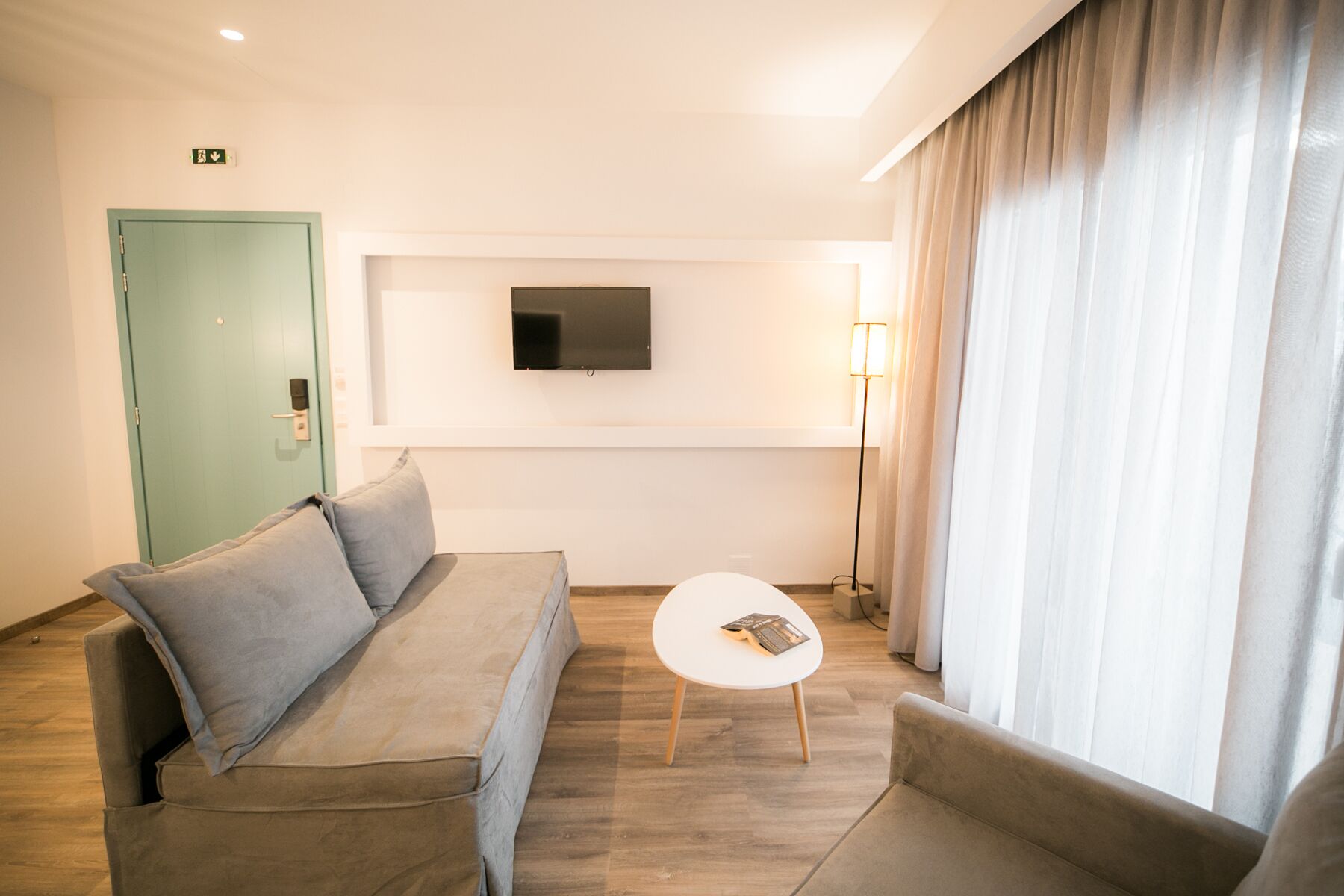 Ein modernes Hotelzimmer mit grauem Sofa, einem kleinen weißen Tisch und einem an der Wand befestigten Fernseher.