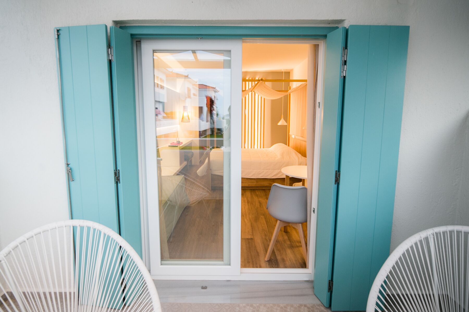 Habitación con puerta corrediza de vidrio que da a una cama y una pequeña mesa, sillas blancas y vistas al exterior.