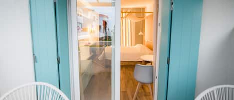 Habitación con puerta corrediza de vidrio que da a una cama y una pequeña mesa, sillas blancas y vistas al exterior.
