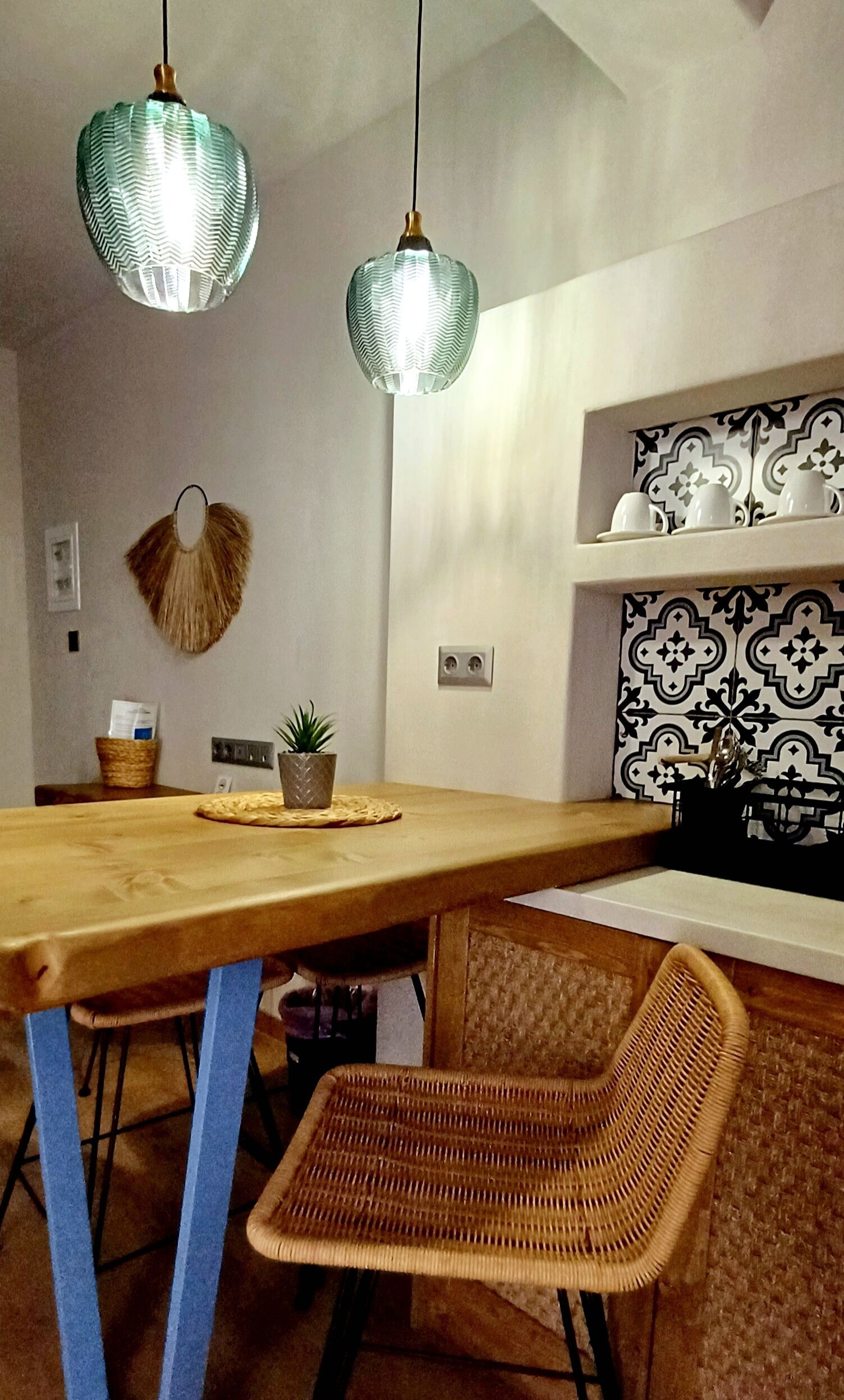 Uma área de jantar com mesa de madeira, duas luminárias pendentes, uma cadeira de vime, um vaso com planta e uma prateleira na parede com objetos decorativos.