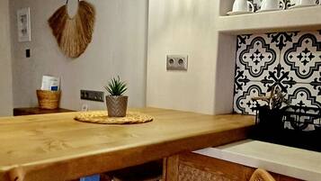 Uma área de jantar com mesa de madeira, duas luminárias pendentes, uma cadeira de vime, um vaso com planta e uma prateleira na parede com objetos decorativos.