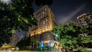 Un edificio alto con varios pisos, una entrada curva y abundante vegetación alrededor.
