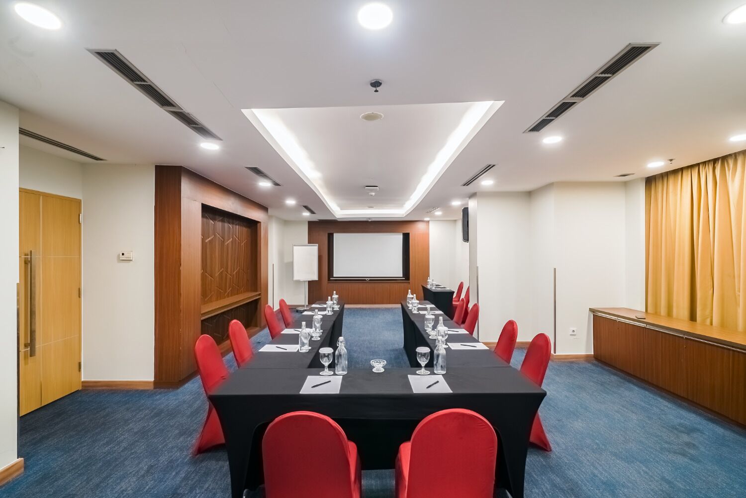 Een vergaderzaal met een lange tafel, rode stoelen, een projectiescherm en een whiteboard.