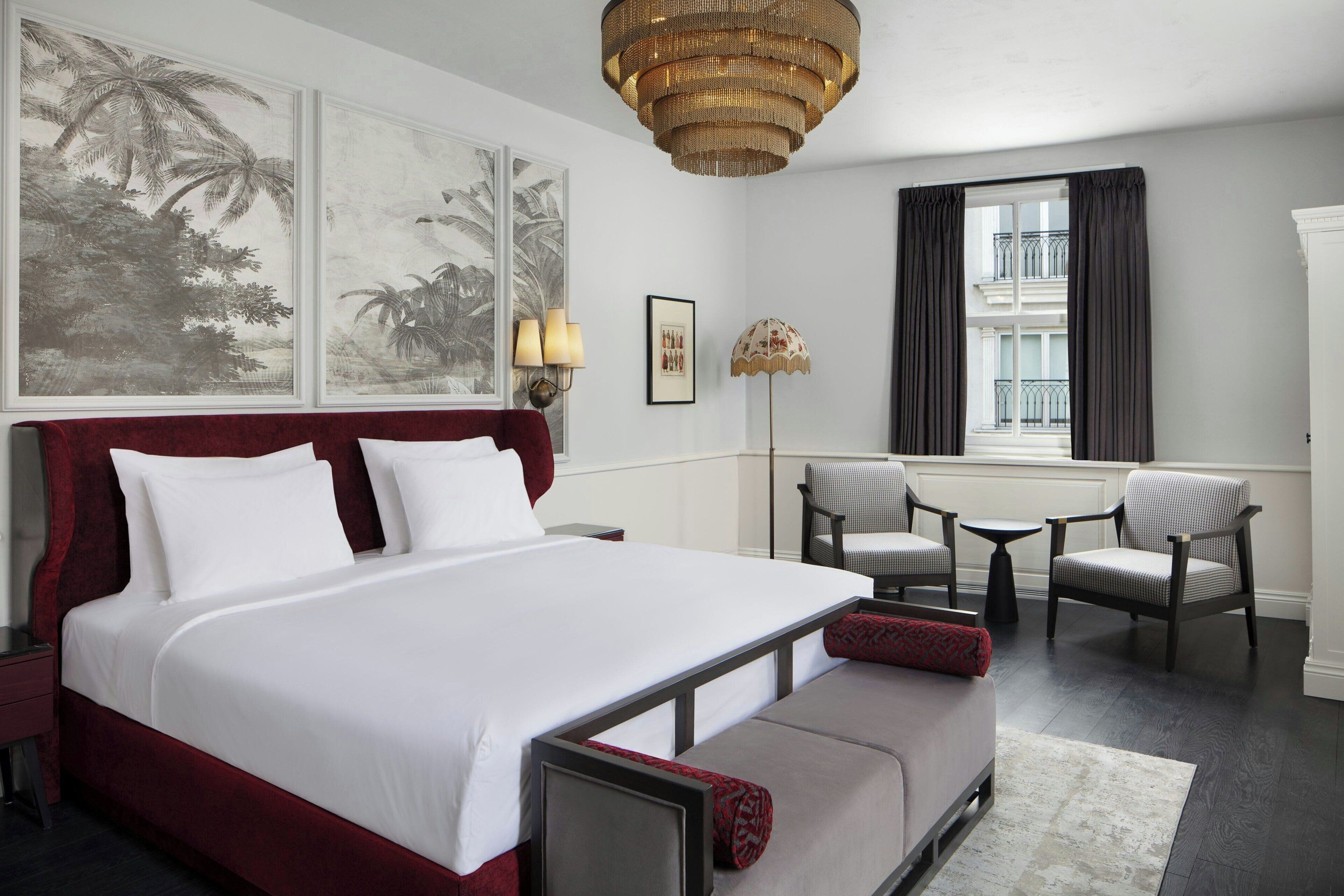 Ein Hotelzimmer mit einem großen Bett, einer Couch, einem Schreibtisch und einem Sessel. Zwei Wandlampen, eine Stehlampe und ein Fenster mit Vorhängen sind vorhanden.