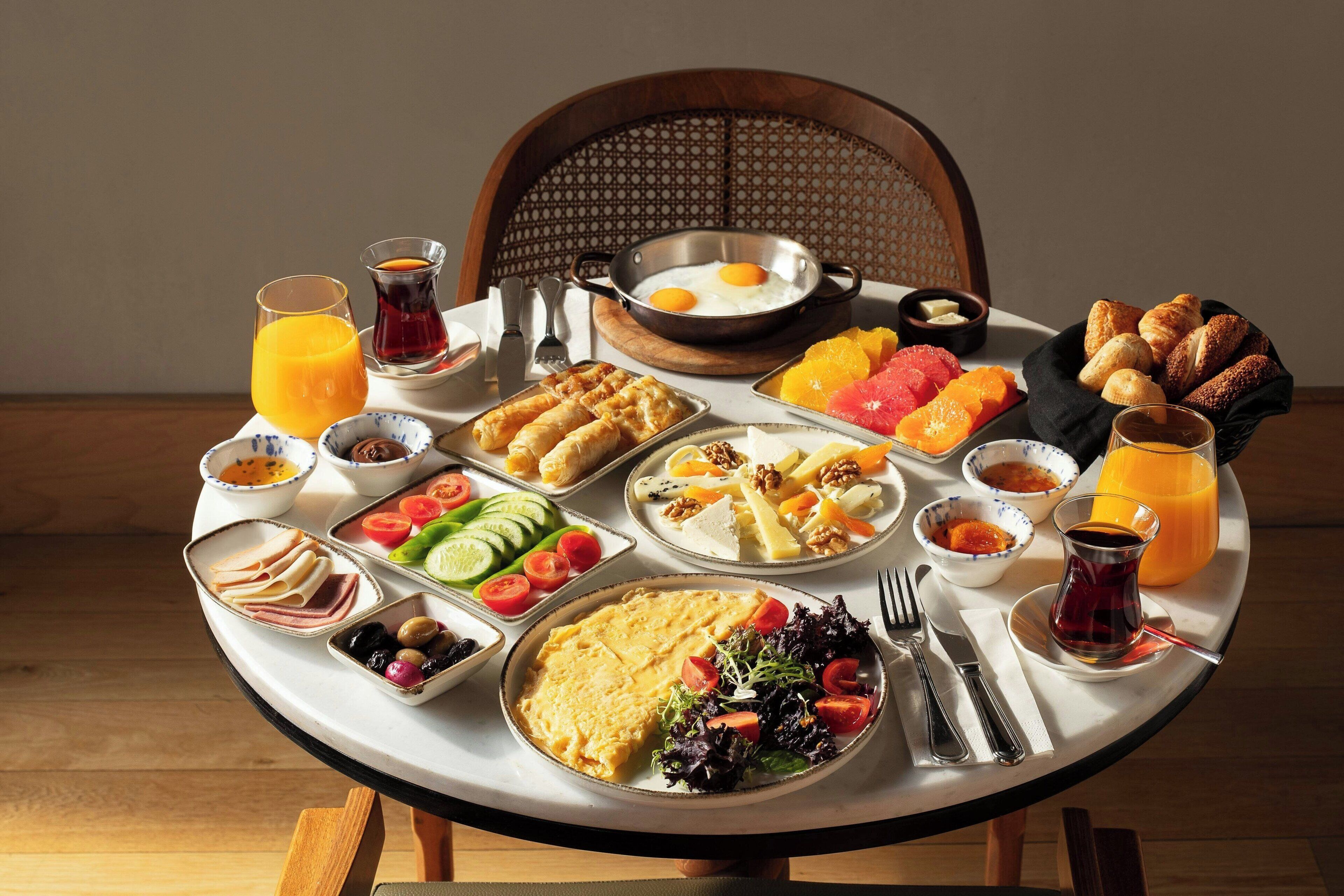 Una mesa de desayuno con diversos platos, entre ellos huevos, bollería, frutas y bebidas.