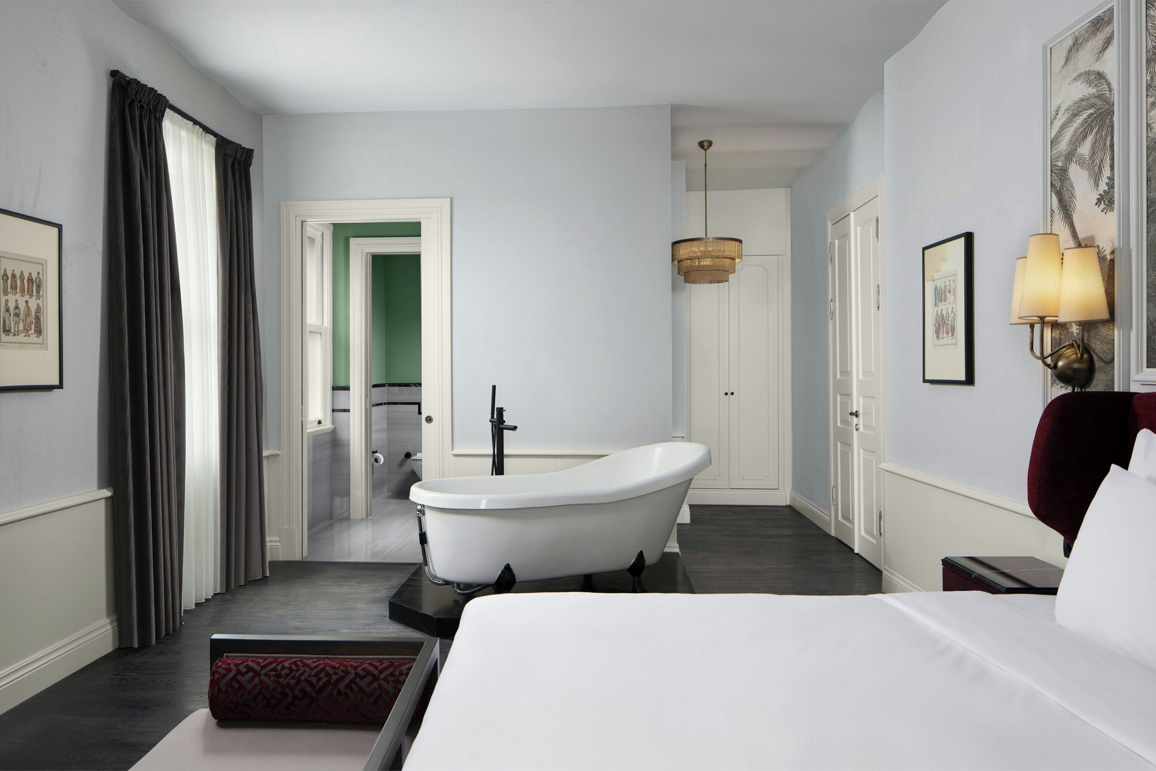 A modern hotel room with a large bed, a freestanding bathtub, a chandelier, and a framed picture on the wall.