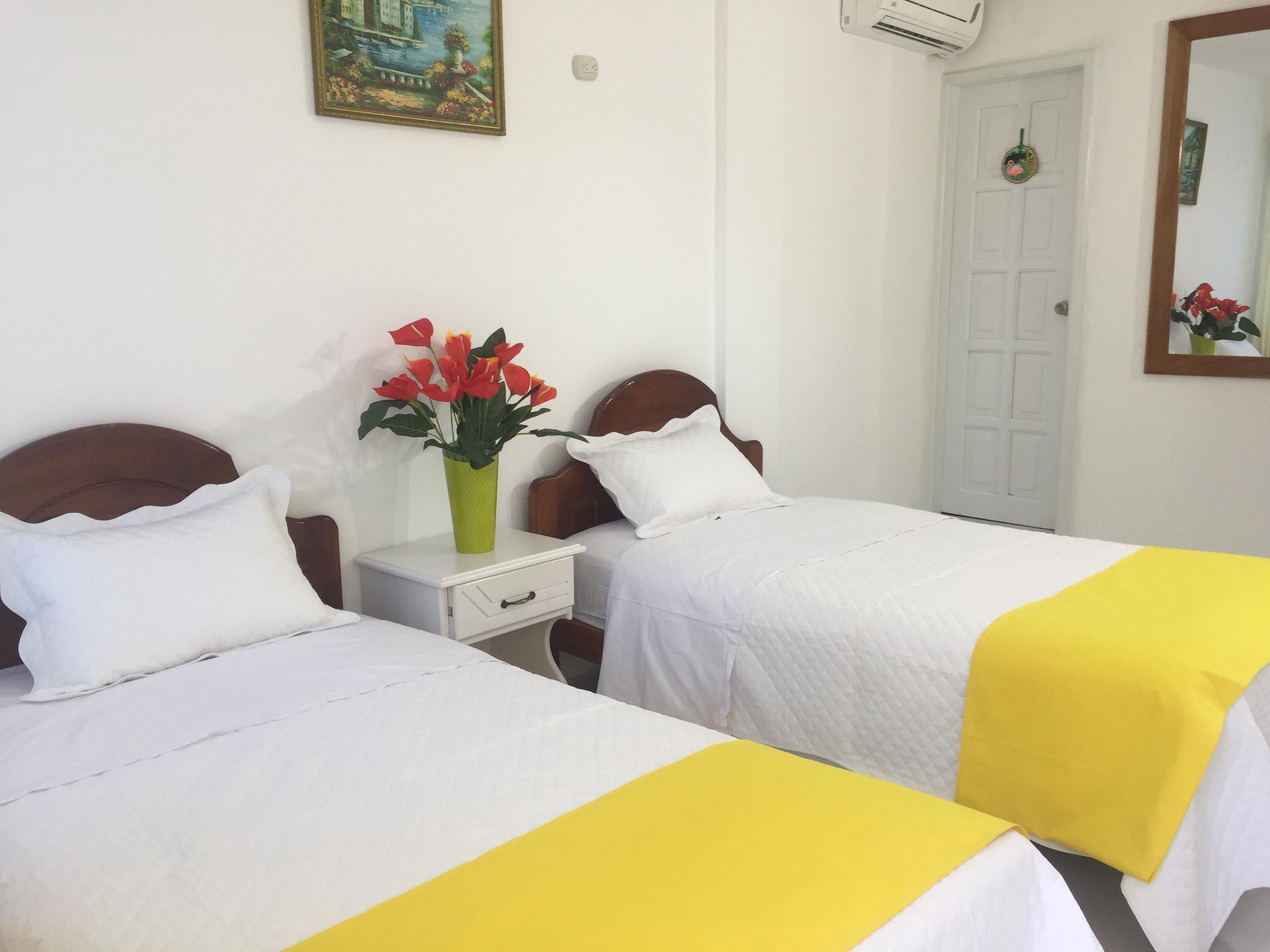 A hotel room with two beds, white bedding, a nightstand with a vase of red flowers, and a painting on the wall.