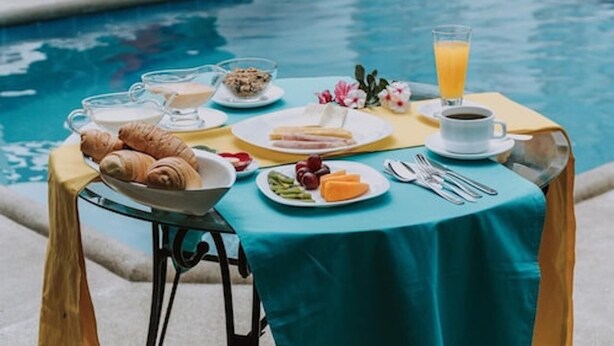 A breakfast setup by a pool with croissants, fruit, cheese, and coffee. - Hostal Loja (Puerto Villamil)