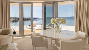 Une pièce moderne avec une table ronde, des chaises blanches, un canapé et une vue sur une étendue d’eau, visible à travers de grandes portes vitrées.