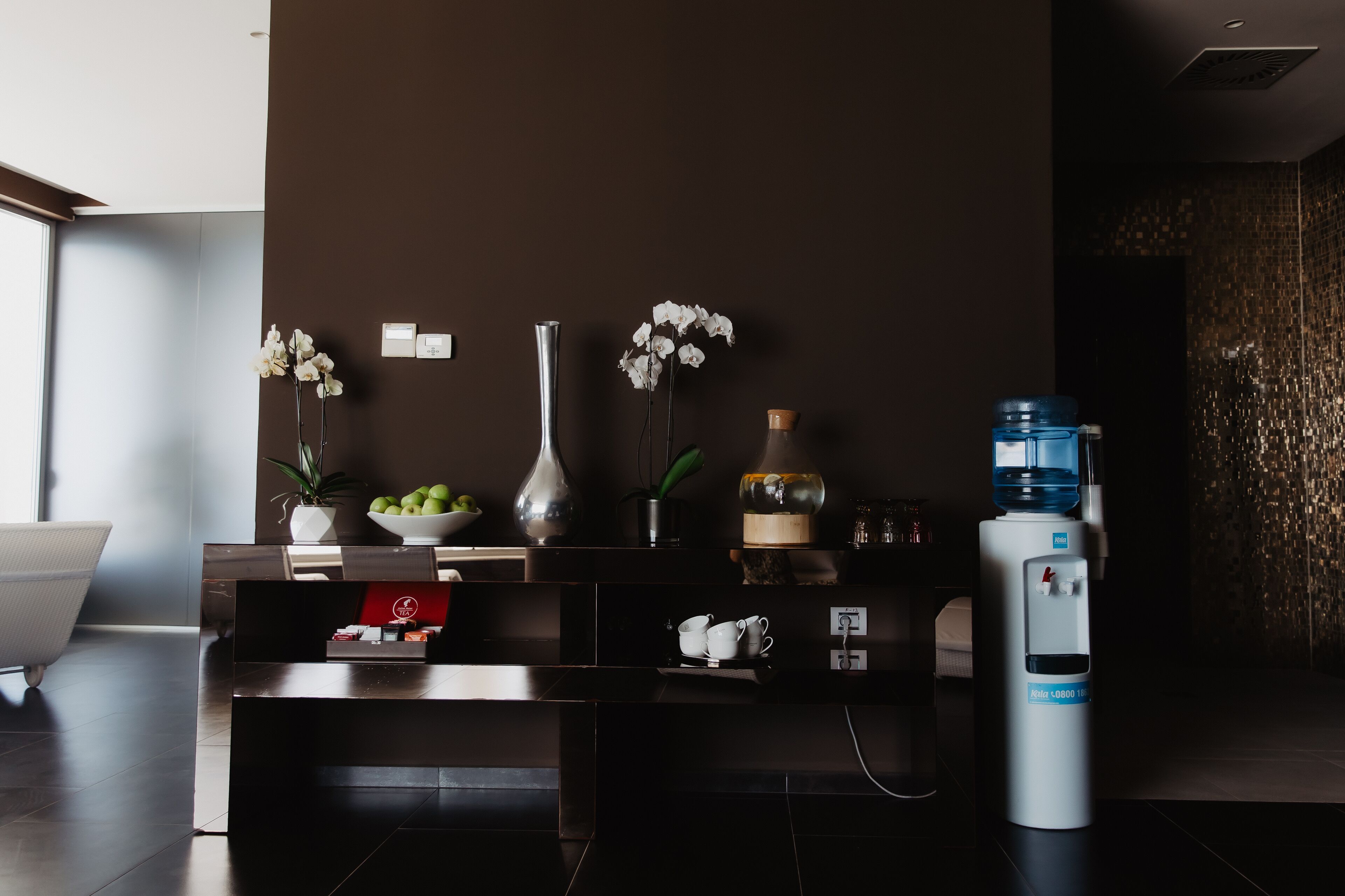 Un área de recepción moderna con una pared marrón oscuro, un dispensador de agua, un jarrón con flores blancas, un frutero y una planta en maceta.