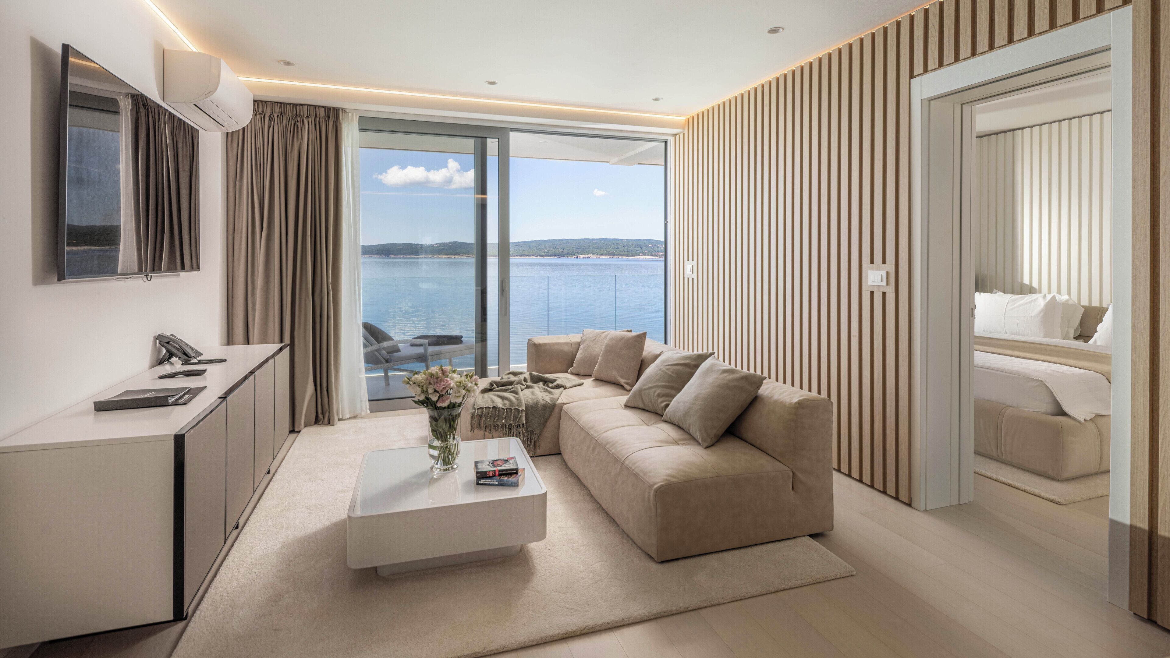 A modern hotel room with a sofa, a coffee table, a desk, and a large window offering a view of the sea.