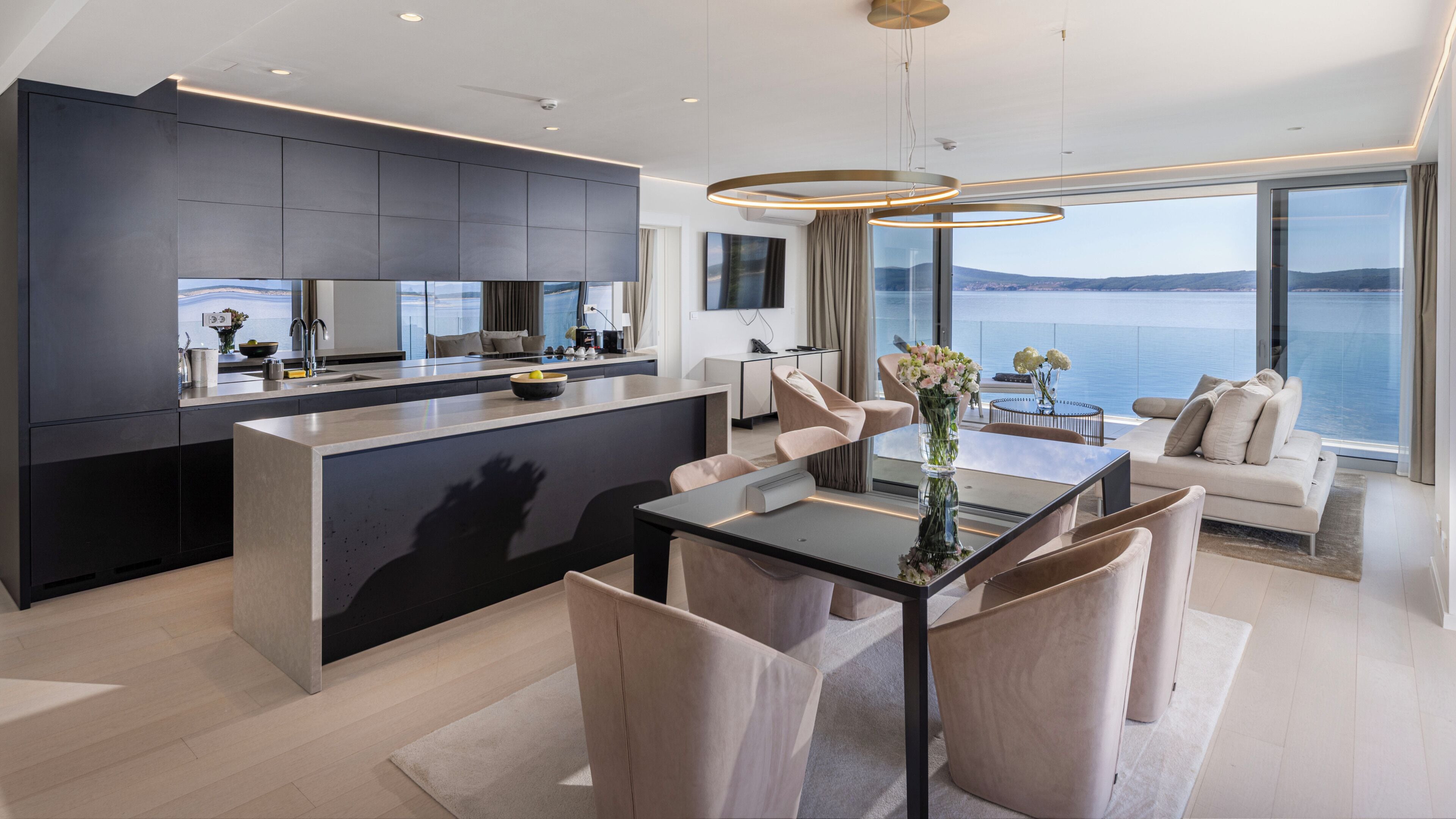 A modern kitchen with a central island, a dining area with a glass table, and a living space with a sofa and a view of the water.
