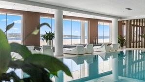 Une piscine intérieure offrant une vue dégagée sur la mer, des chaises longues blanches et de grandes fenêtres.