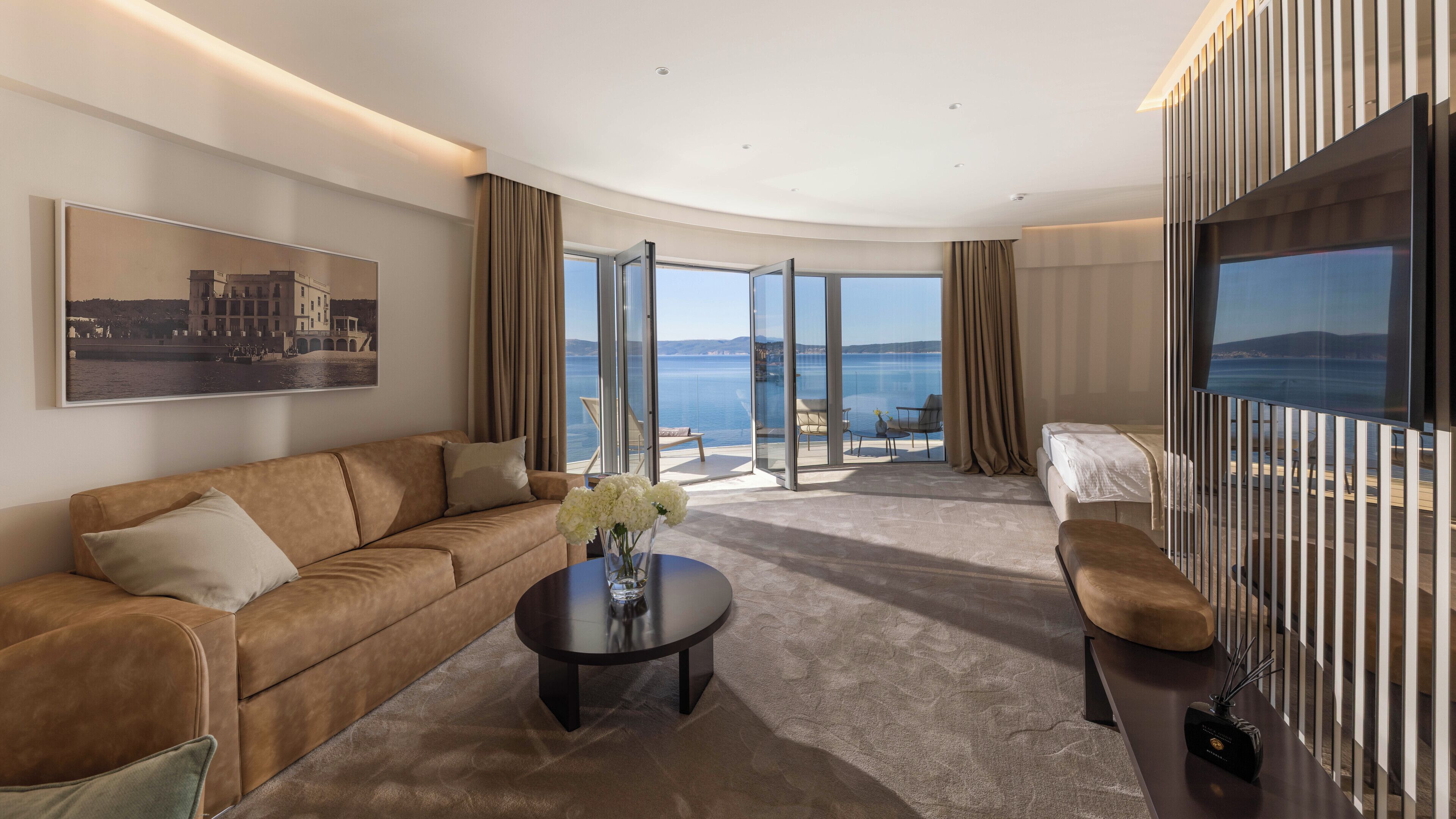 A modern hotel room with a curved ceiling, a large window with a view of the sea, a sofa, a coffee table, and a TV.