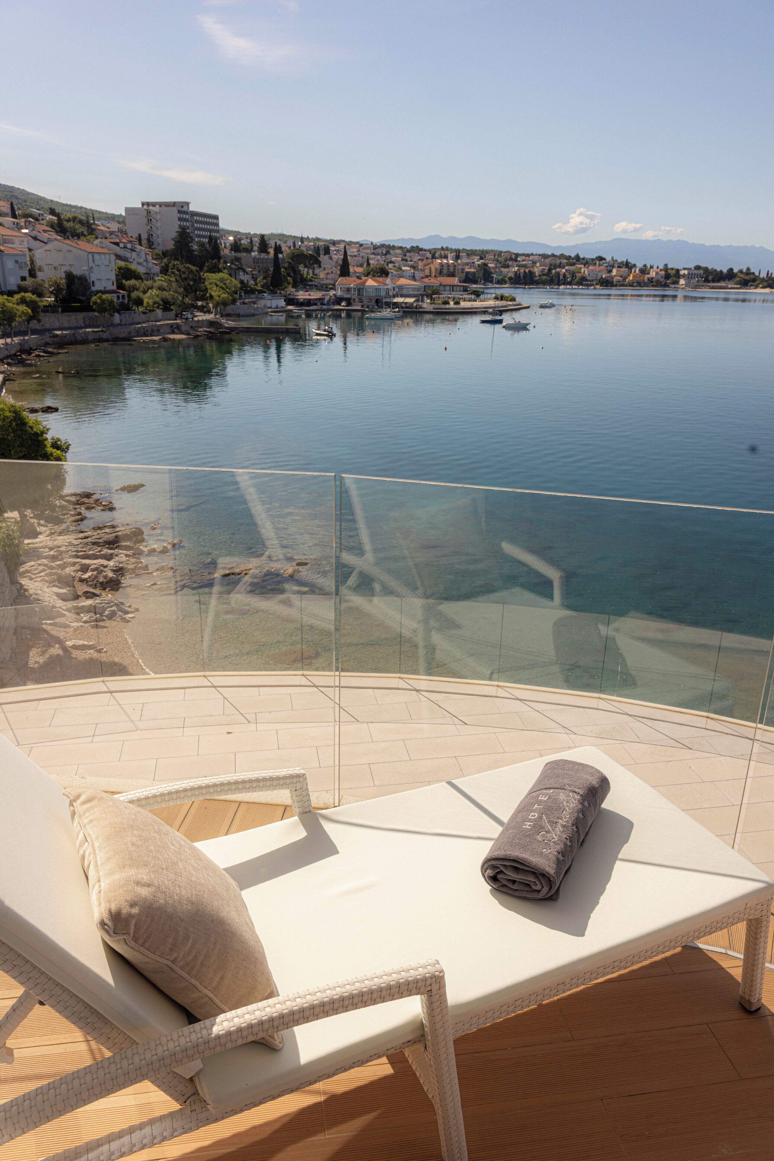 A lounge chair with a towel, a glass balustrade, and a view of a calm lake with buildings in the background.