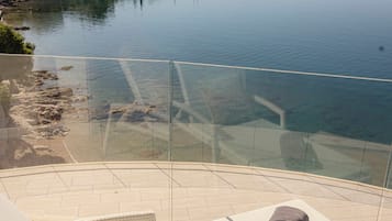 A lounge chair with a towel, a glass balustrade, and a view of a calm lake with buildings in the background.