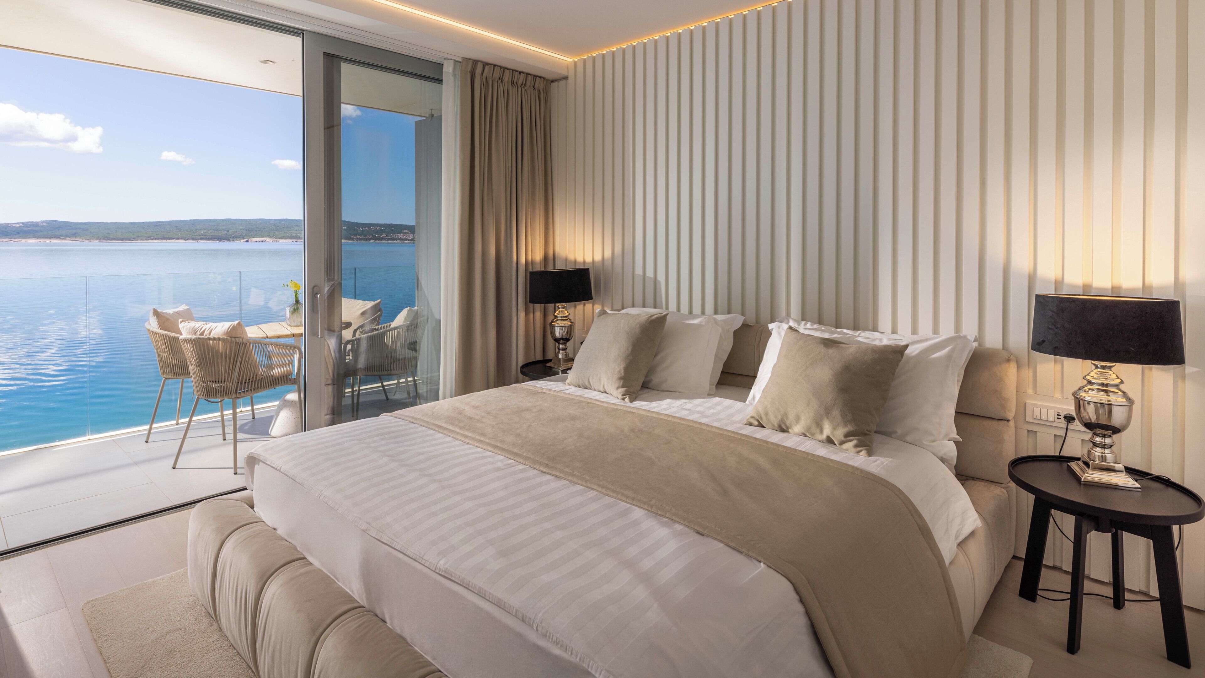 A hotel room with a large bed, two bedside lamps, a nightstand, a glass door leading to a balcony with a view of the sea.