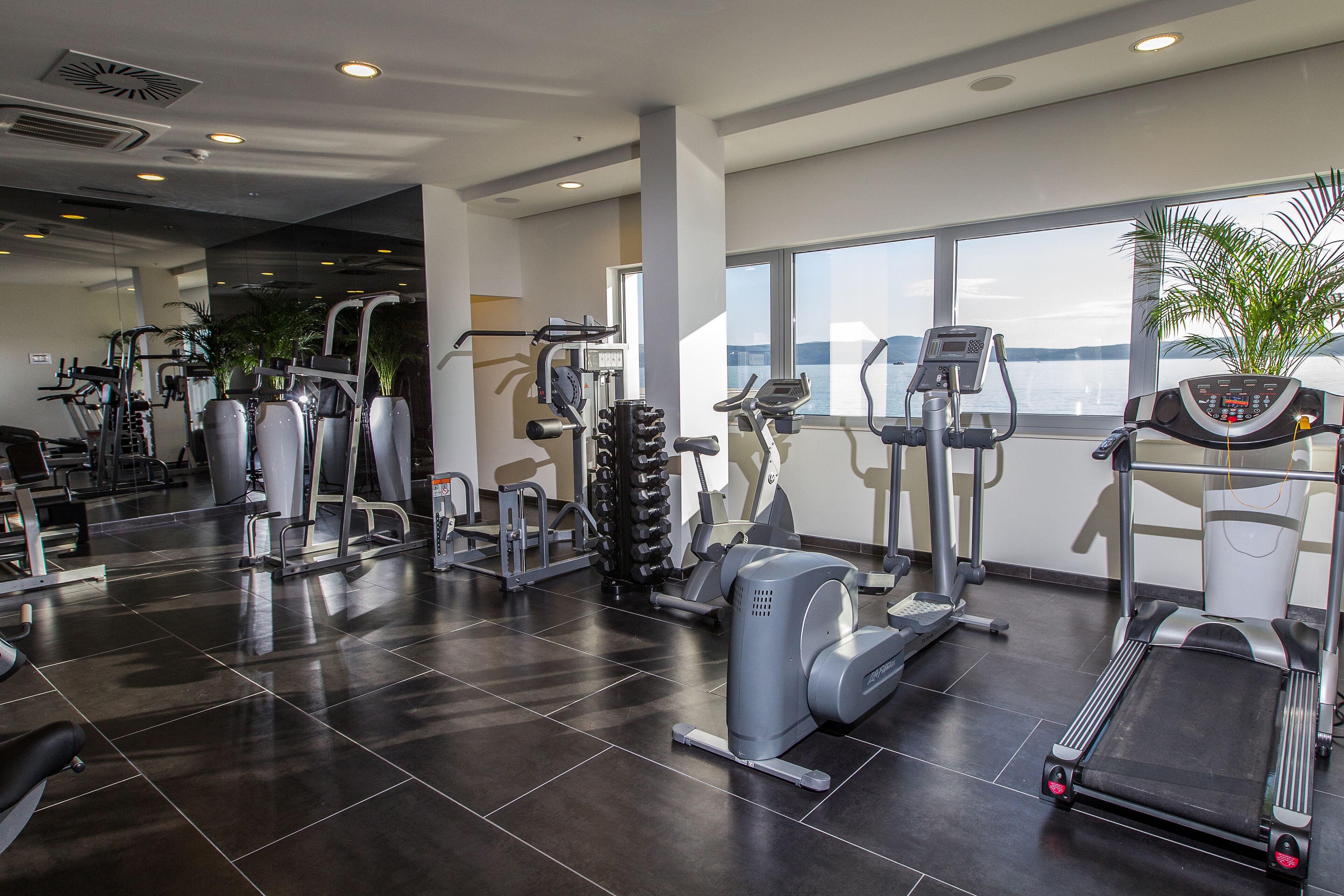 A modern gym with various exercise equipment, large windows, and a view of the water.