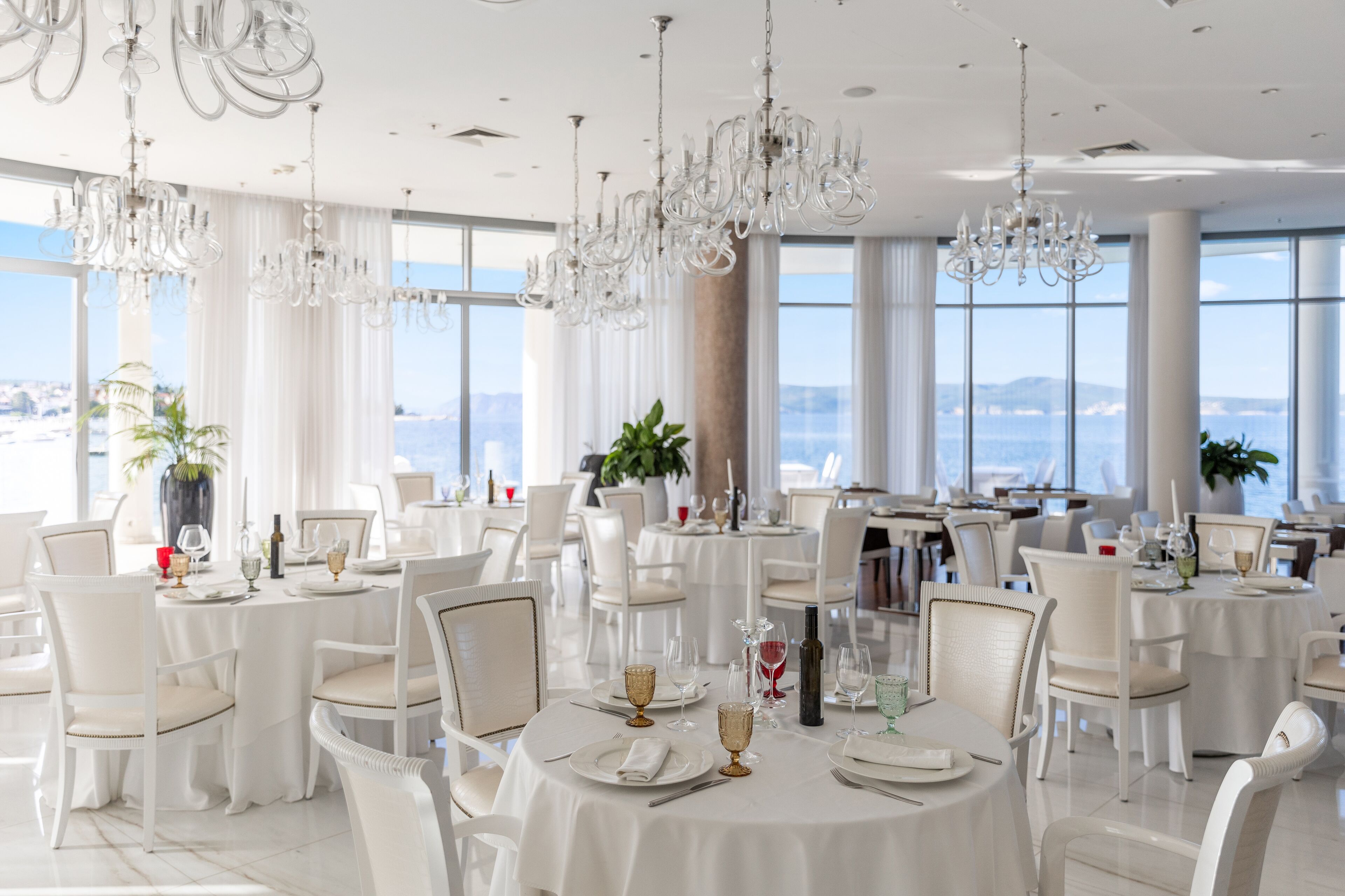 A dining area with round tables set for a meal, white chairs, and chandeliers.