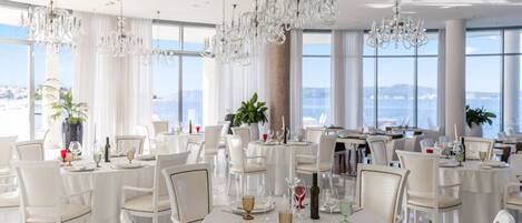 A dining area with round tables set for a meal, white chairs, and chandeliers.