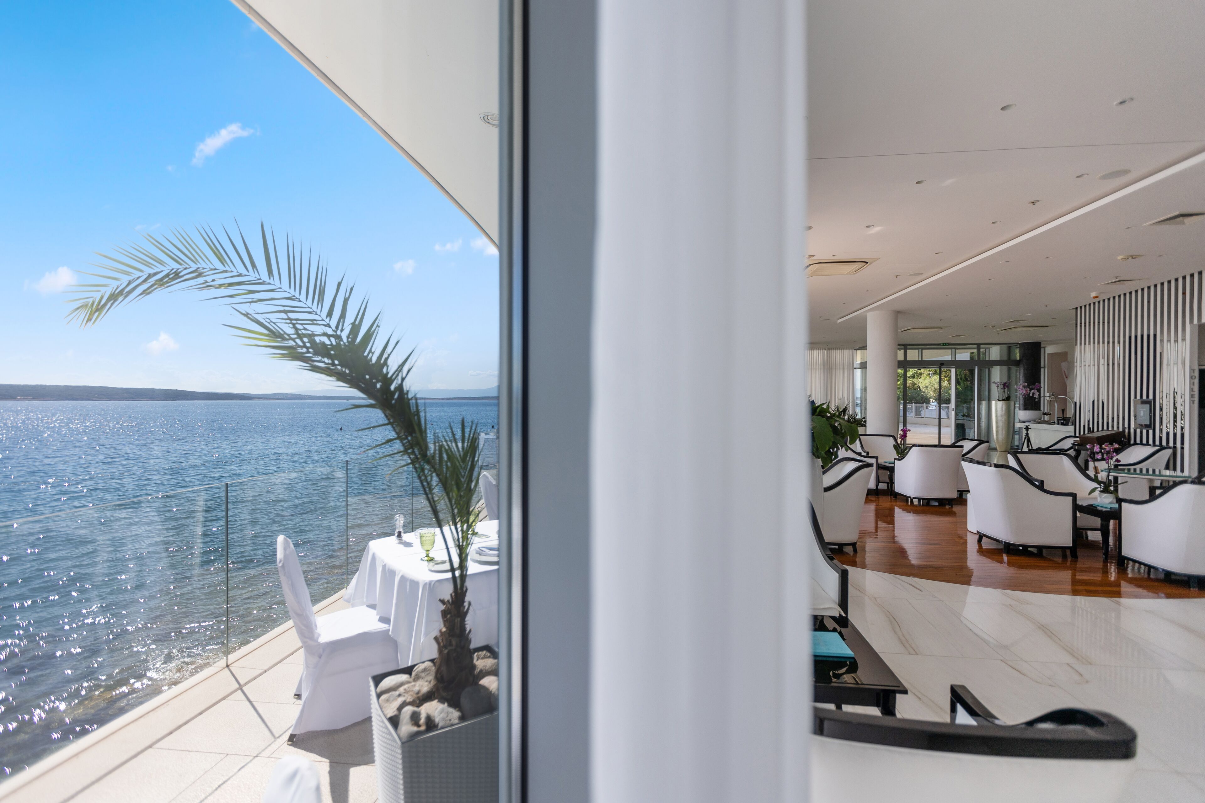 Habitación de hotel moderna con un balcón grande con vistas al mar, amueblada con sillas y mesas blancas, e interior espacioso con techo alto y decoración moderna.