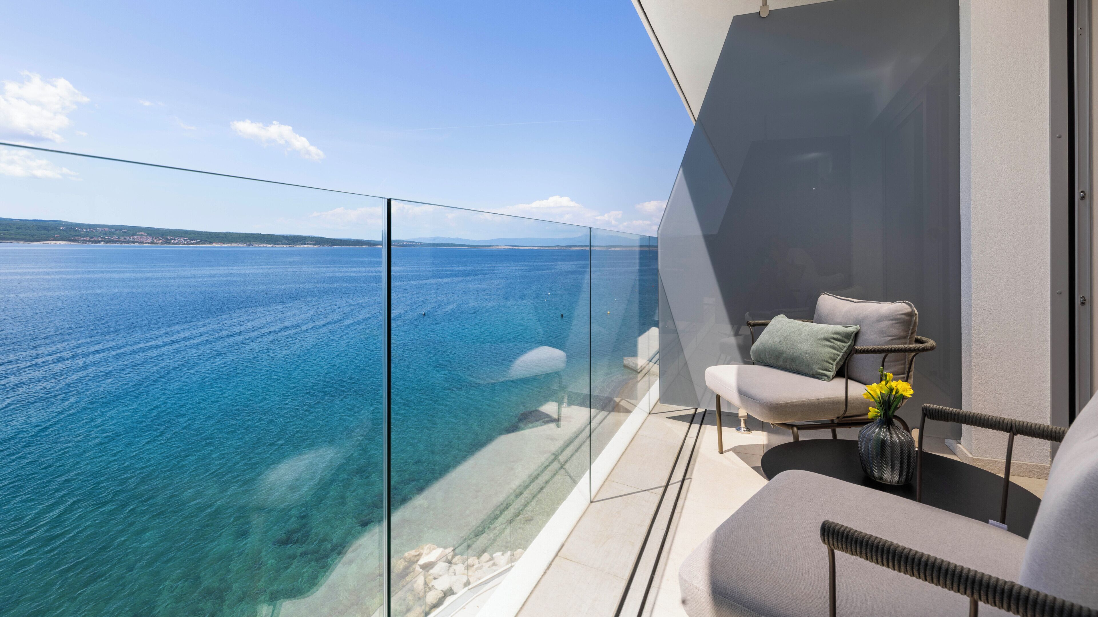 A balcony with a clear view of the sea, a chair, and a small table with a vase of flowers.