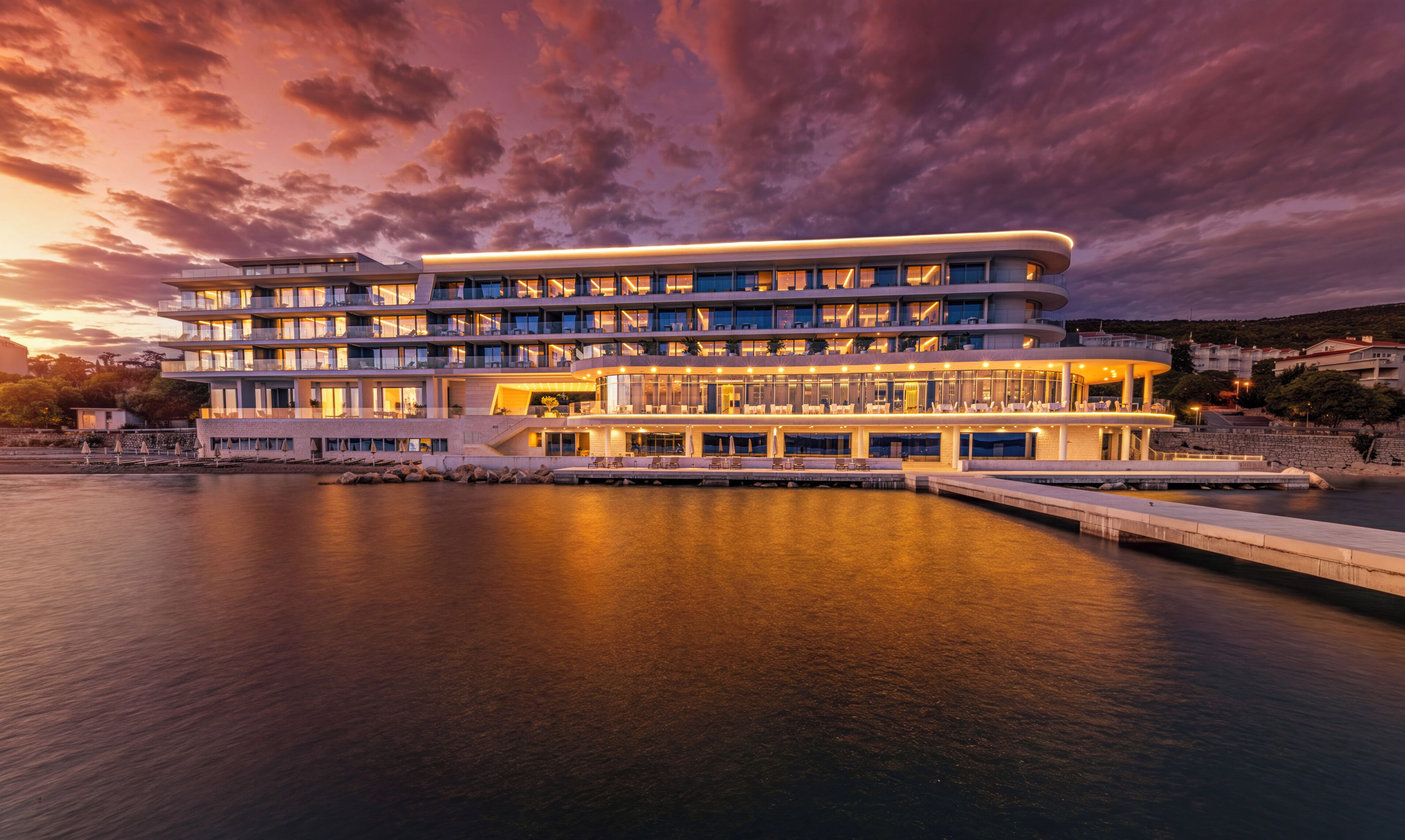 Un hôtel à plusieurs étages doté de balcons, offrant une vue sur le front de mer et comprenant un quai s’avançant dans l’eau.