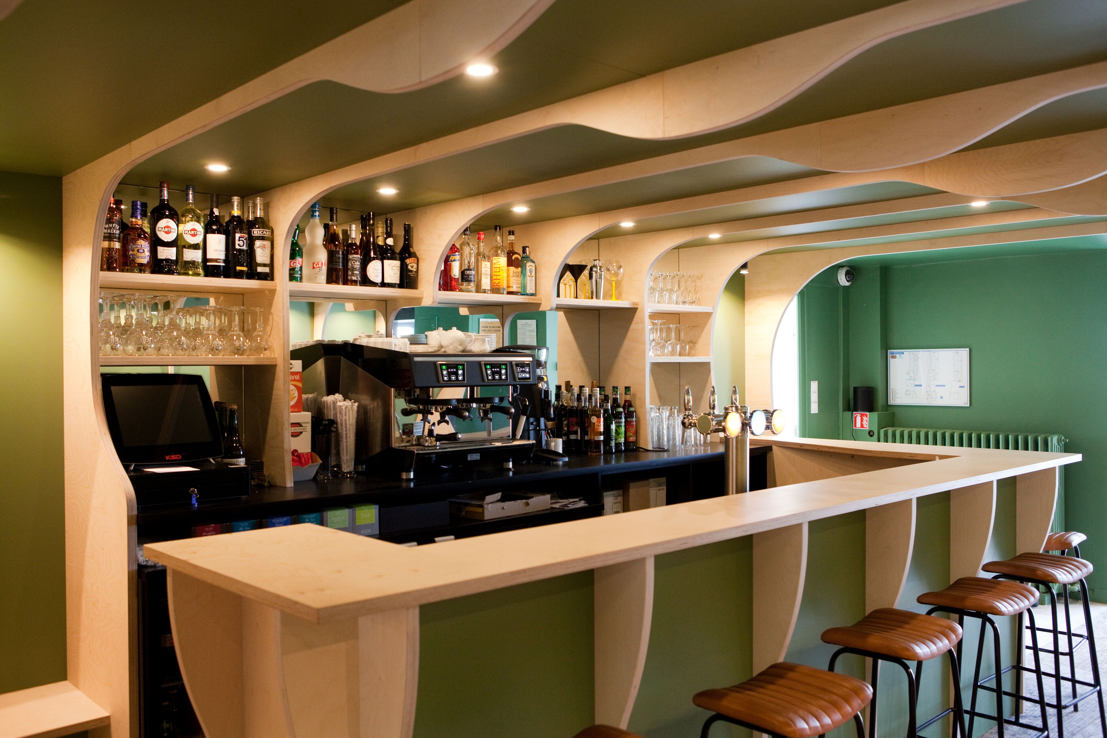 A bar area with a long counter, stools, shelves with bottles, glasses, and a cash register.