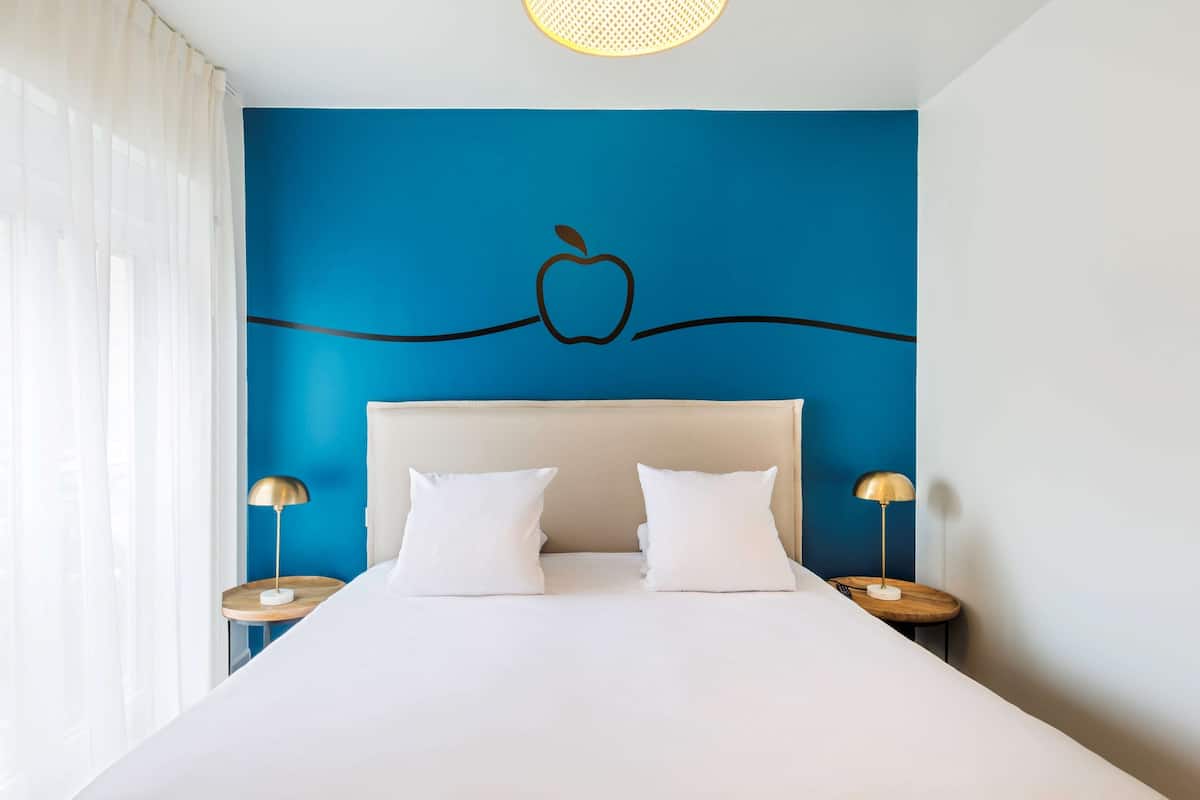 Habitación de hotel con una cama grande, dos mesitas de noche y una pared azul con el logo de Apple.