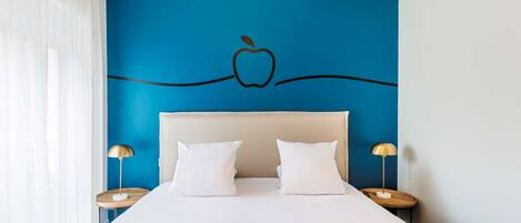 Habitación de hotel con una cama grande, dos mesitas de noche y una pared azul con el logo de Apple.