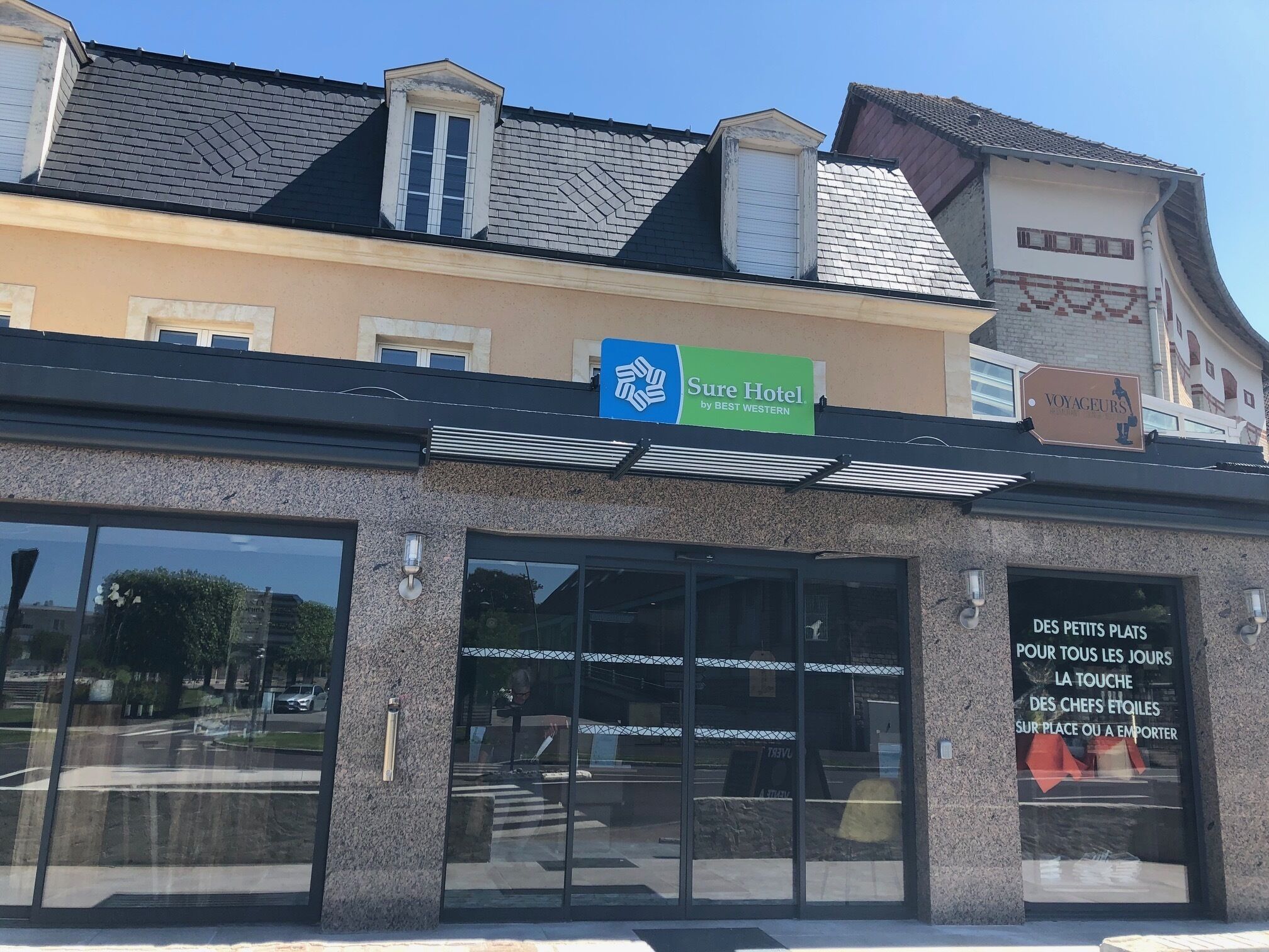 Un hotel con un cartel que dice 'Sure Hotel by BEST WESTERN' y un escaparate que anuncia servicio de pequeñas porciones diarias: 'DES PETITS PLATS POUR TOUS LES JOURS'.
