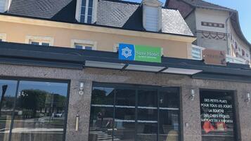Un hotel con un cartel que dice 'Sure Hotel by BEST WESTERN' y un escaparate que anuncia servicio de pequeñas porciones diarias: 'DES PETITS PLATS POUR TOUS LES JOURS'.