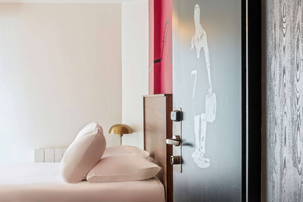 Un dormitorio con cama, almohadas, una mesita de noche con lámpara, una puerta con número y un grafiti en la pared.