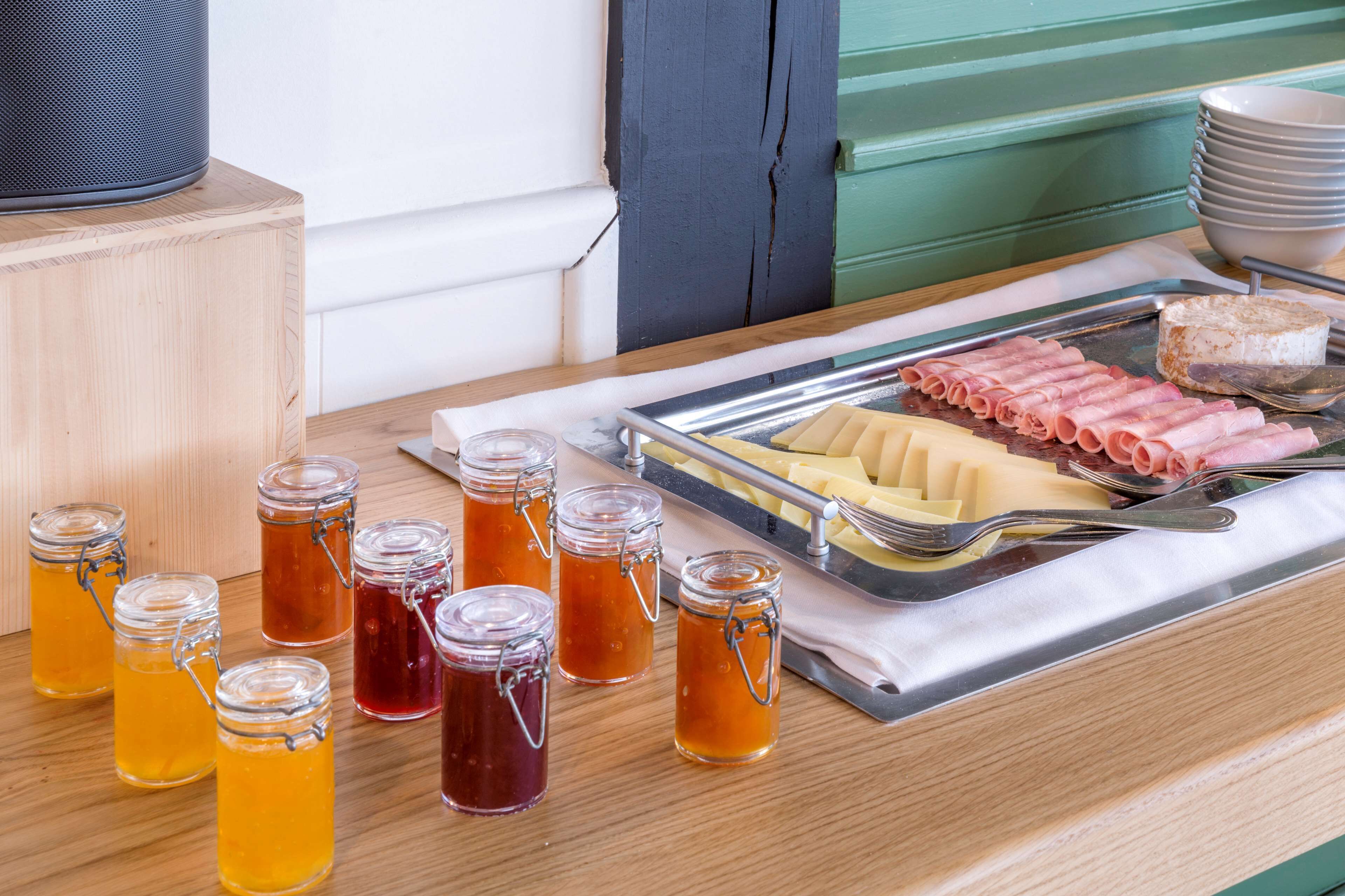 A breakfast tray with ham, cheese, and a cake, accompanied by several jars of jam and honey on a wooden table.