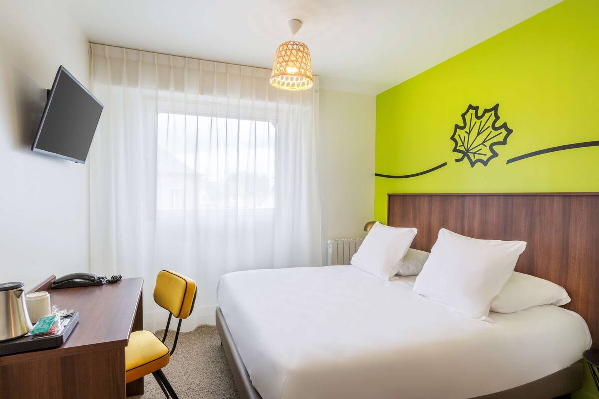 Una habitación de hotel con una cama, un escritorio con una silla, un televisor montado en la pared y una ventana con cortinas translúcidas.