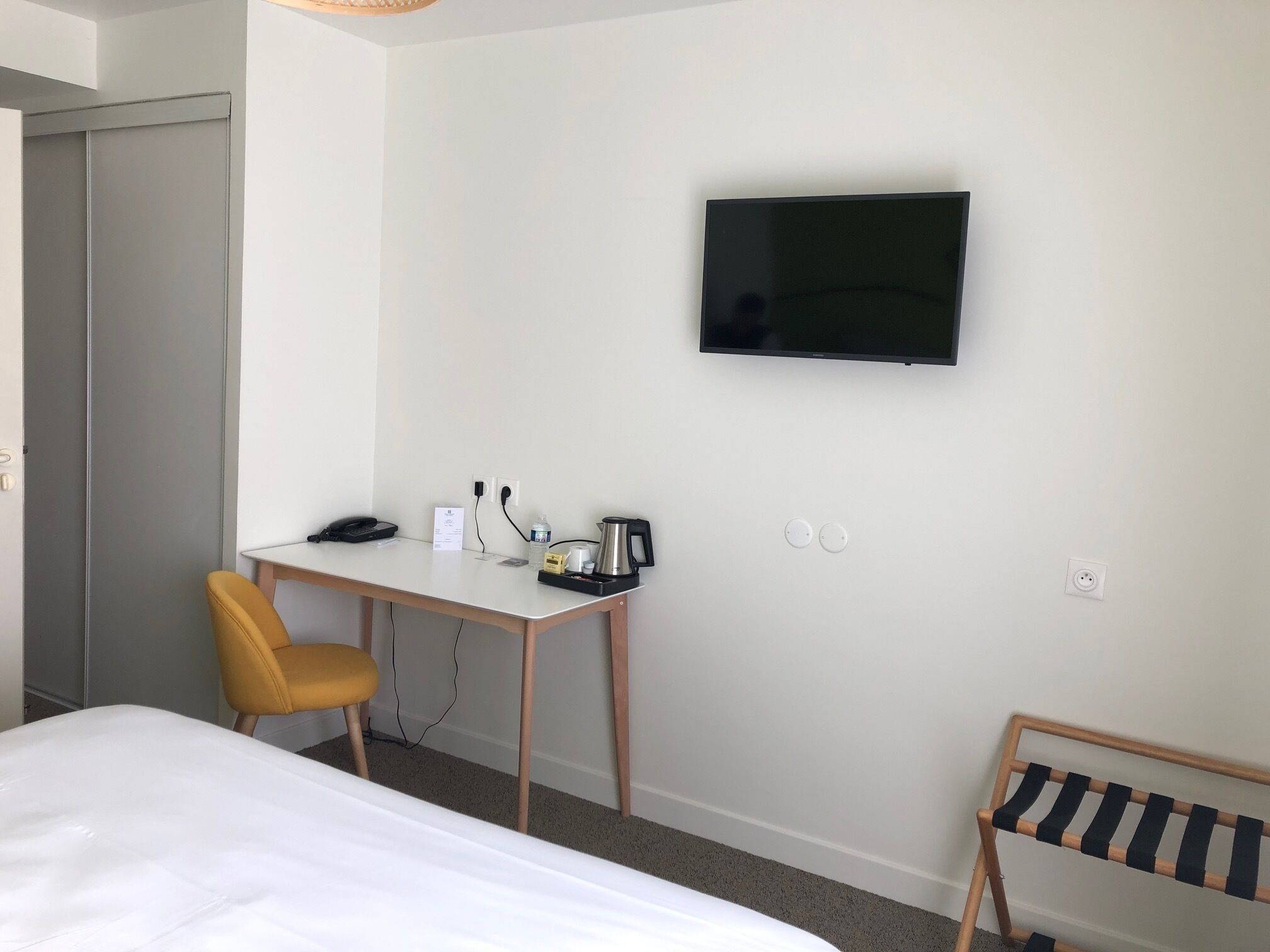 Ein Hotelzimmer mit einem Bett, einem Schreibtisch mit Stuhl, einem Fernseher und einem kleinen Holzregal.