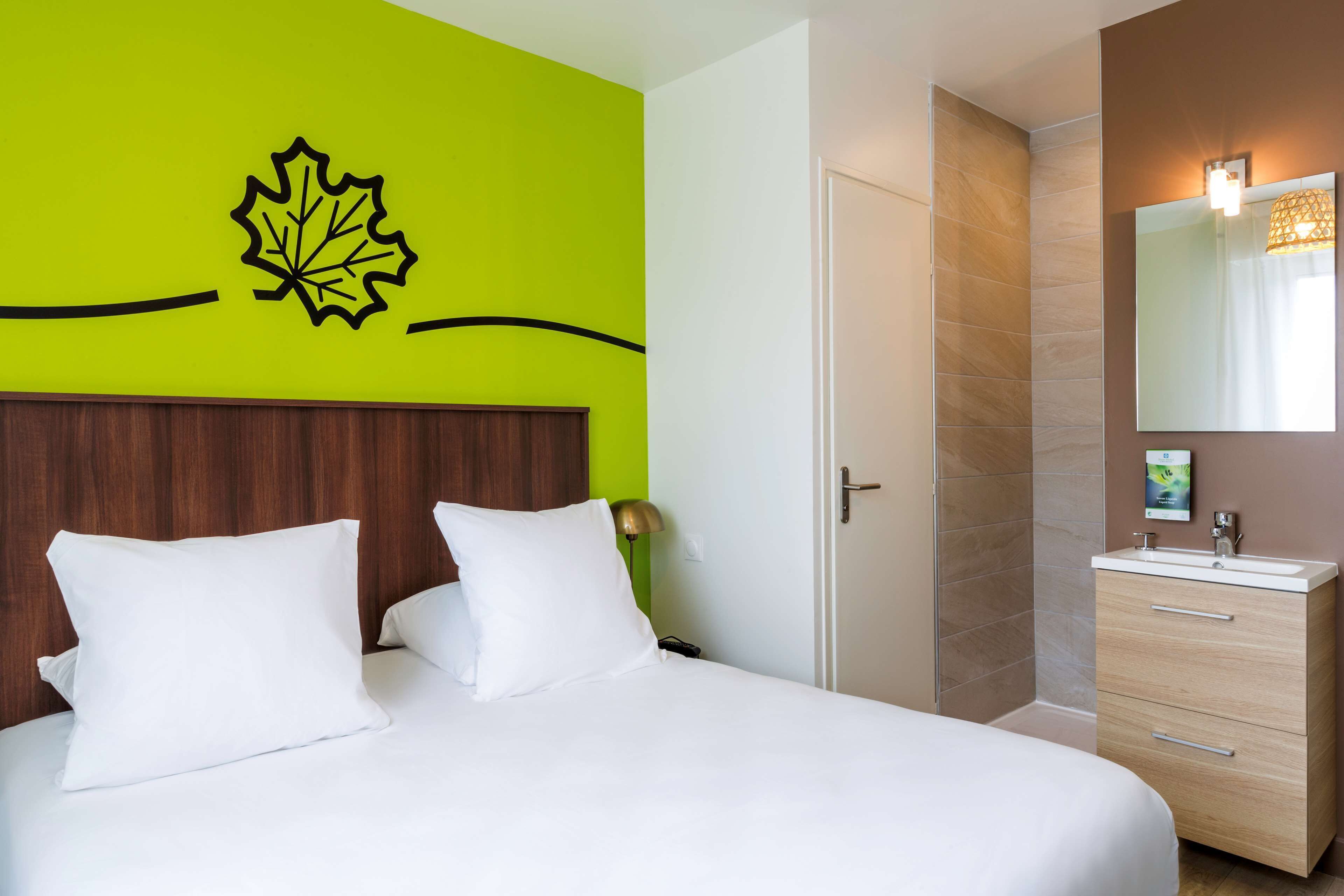 Una habitación de hotel con una cama, cabecera de madera, pared verde brillante con un adhesivo de hoja, baño con espejo y lavamanos, y una puerta.