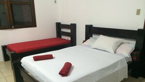 A single bed with white bedding and two red towels, a smaller bed with a red blanket, wooden headboards, and a window with a wooden frame.
