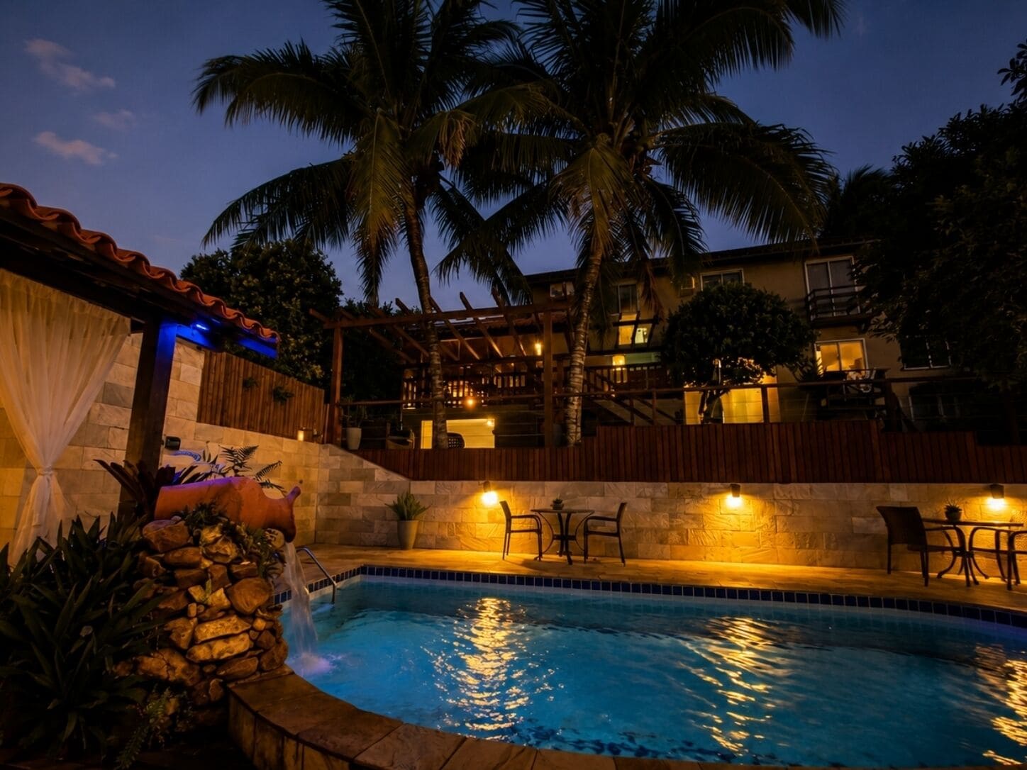A swimming pool with a stone wall, palm trees, and a building with balconies in the background.