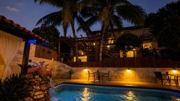 A swimming pool with a stone wall, palm trees, and a building with balconies in the background.