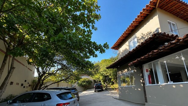 A residential street with parked cars, a building with a tiled roof, and a tree with green leaves. - Calador Búzios Hotel (Búzios)