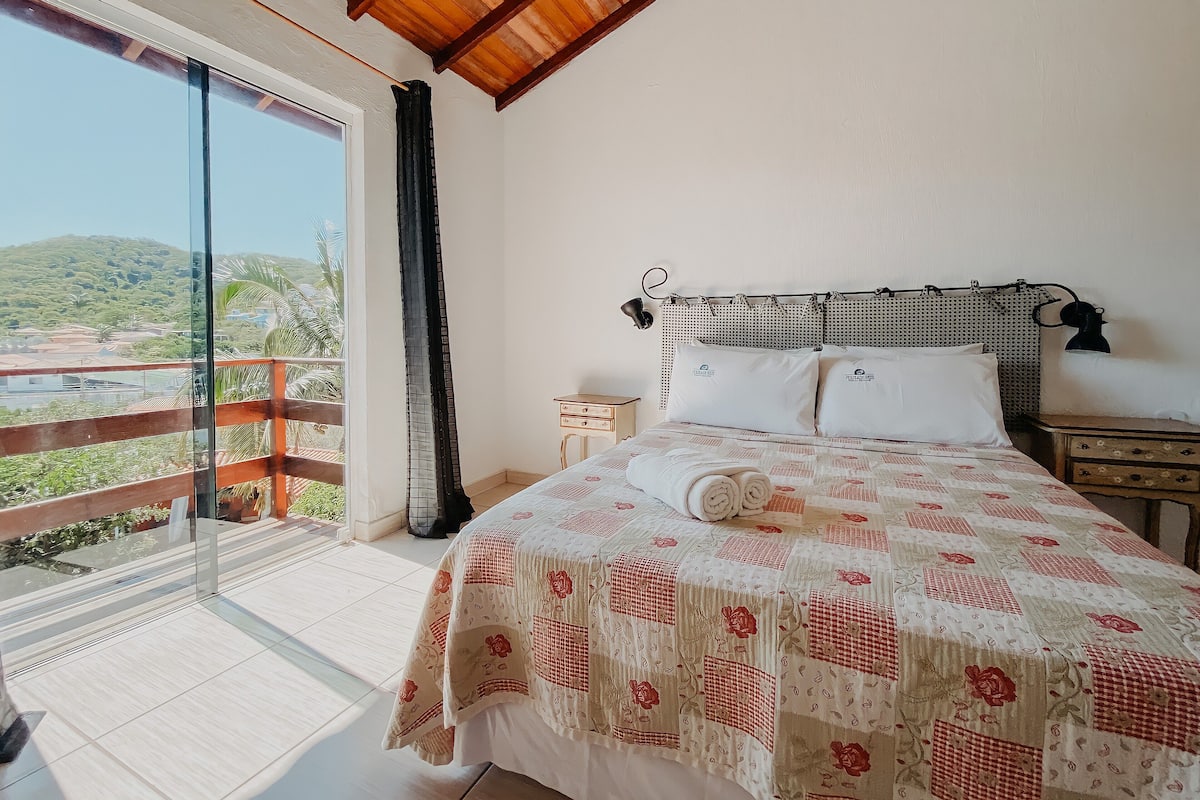 Quarto com uma cama grande, cabeceira de madeira, mesa de cabeceira e vista para uma varanda com guarda-corpo.