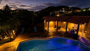 A swimming pool with a tiled deck, a covered patio area with chairs, and a view of a hillside with buildings. - Calador Búzios Hotel (Búzios)